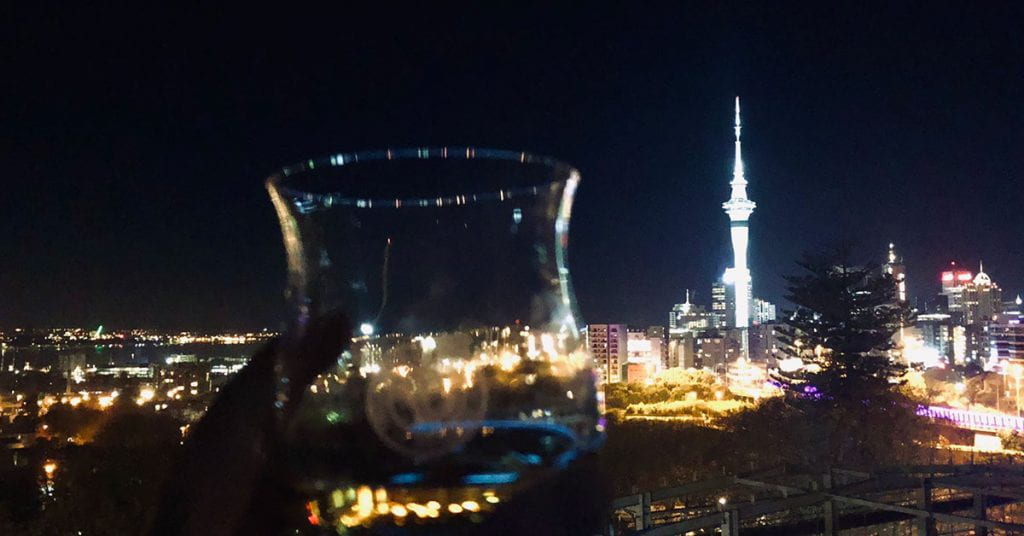 A glass of single malt whiskey held up against a background of the Auckland cityscape at night, including the Sky Tower