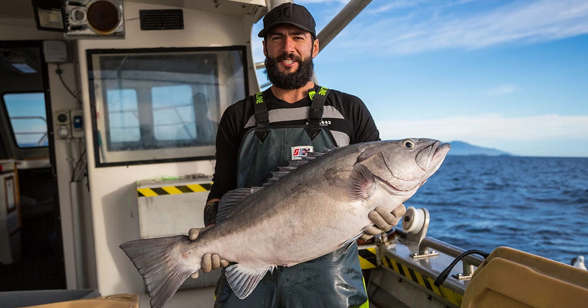 A fisher (Nate Smith) smiling and holding a large fish on a boat