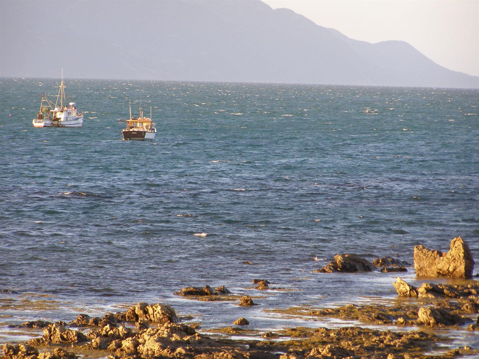 Two fishing vessels near the coast with the Kaikōura mountain in the background, and a rocky shore in the foreground