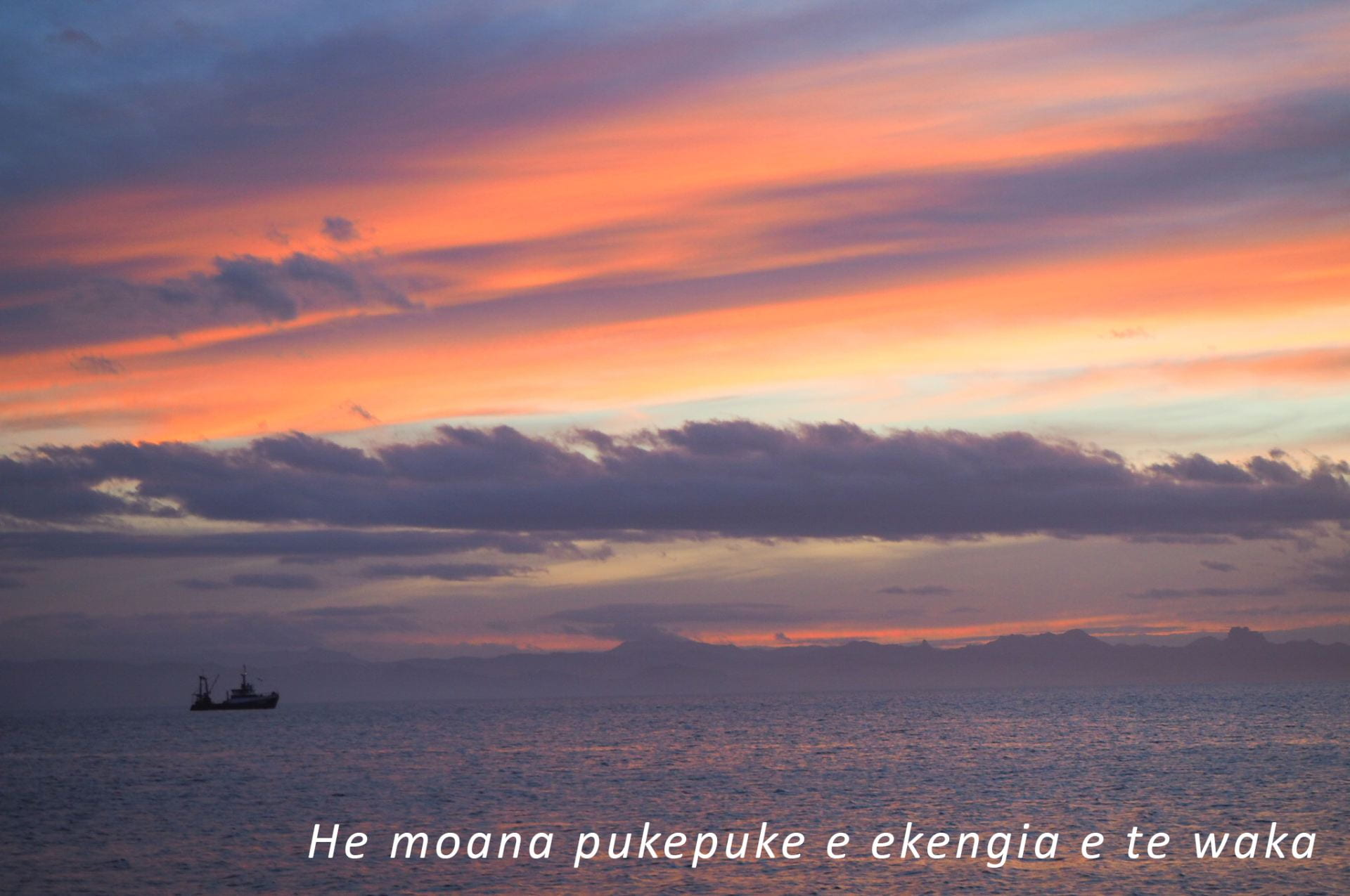 A fishing vessel at sunset off the coast of the Coromandel with the words 'He moana pukepuke e ekengia e te waka'