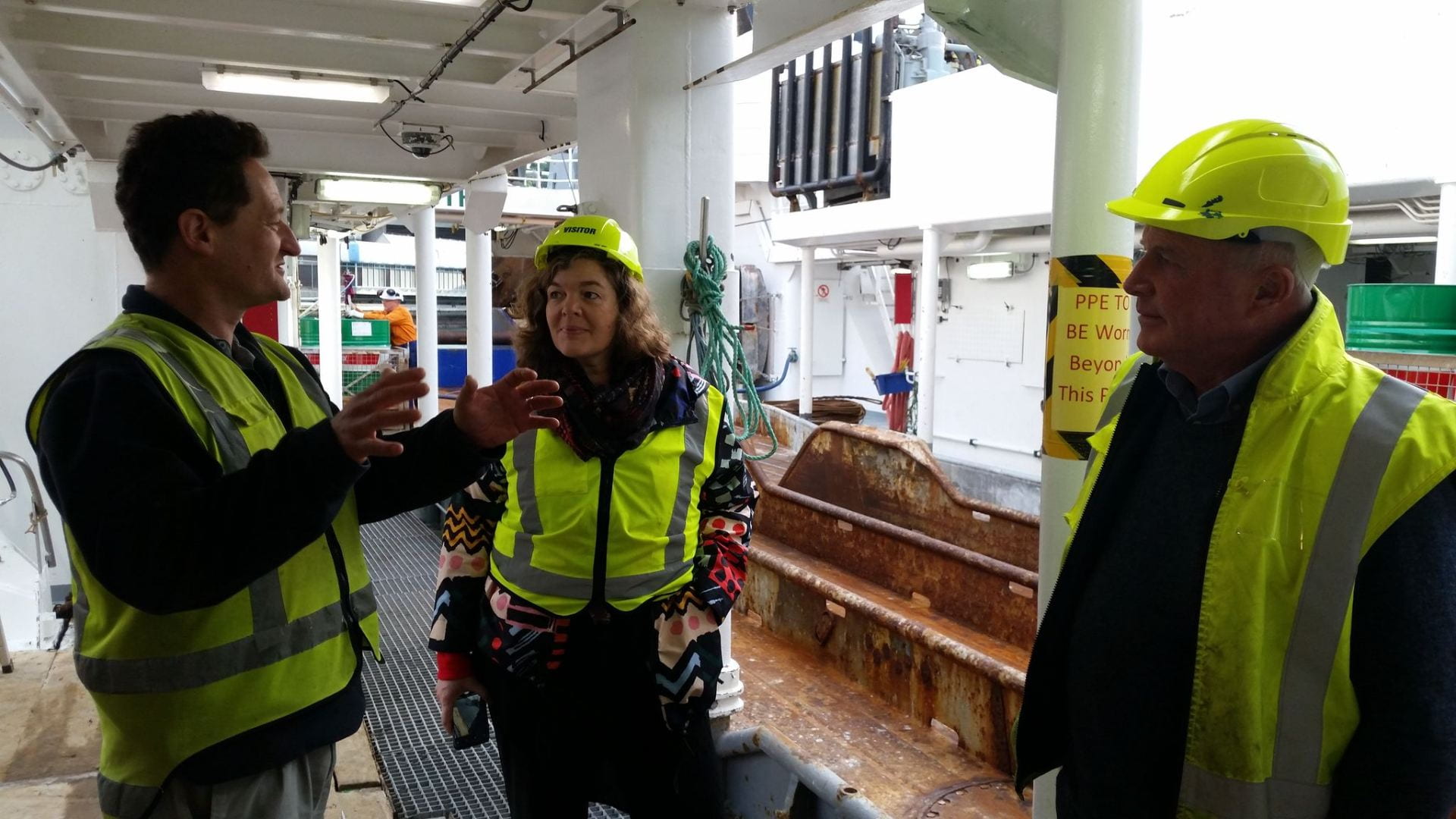 Juliet, wearing a fluoro yellow hard hat and vest, listens to two men (NIWA researchers, also in yellow vests) onboard the RV Tangaroa.