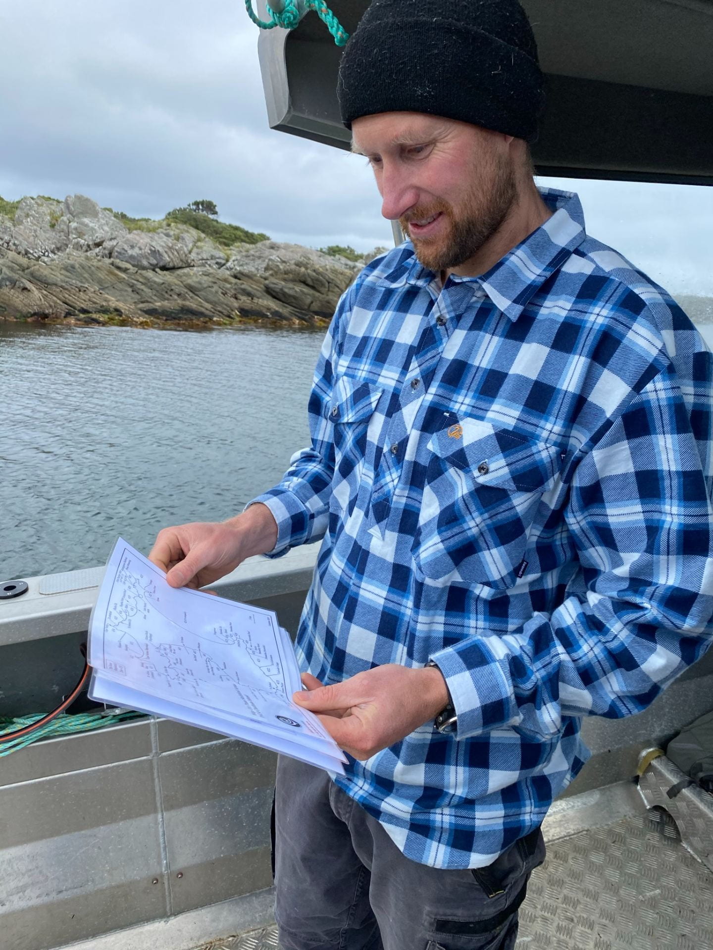 A man in a blue-and-white flannel shirt and a beanie is standing on a boat, holding a map in his hands and smiling. The background is grey sea and sky, with craggy rocks.