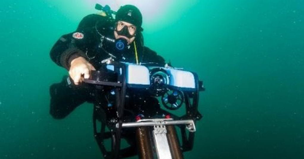 Diver in deep green water holding stereo-camera in two hands, sitting slightly above camera