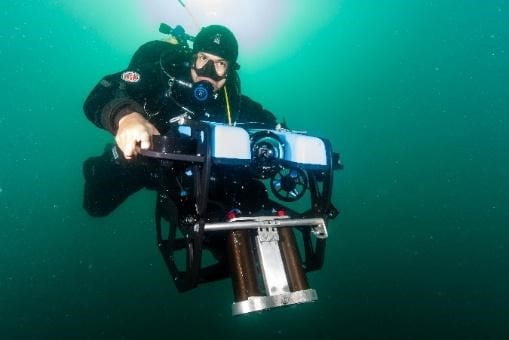 A diver in deep green water is above the camera looking down holding a large stereo-camera in both hands