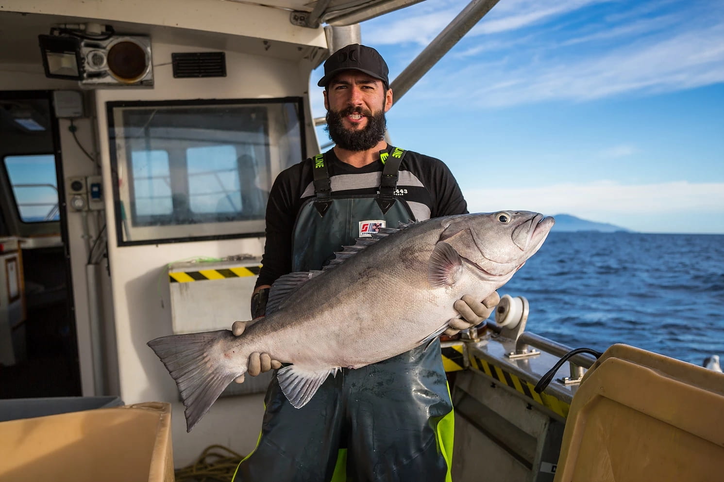 A smiling man with a beard and a cap stands on the deck of a fishing vessel, holding a large silver-grey fish