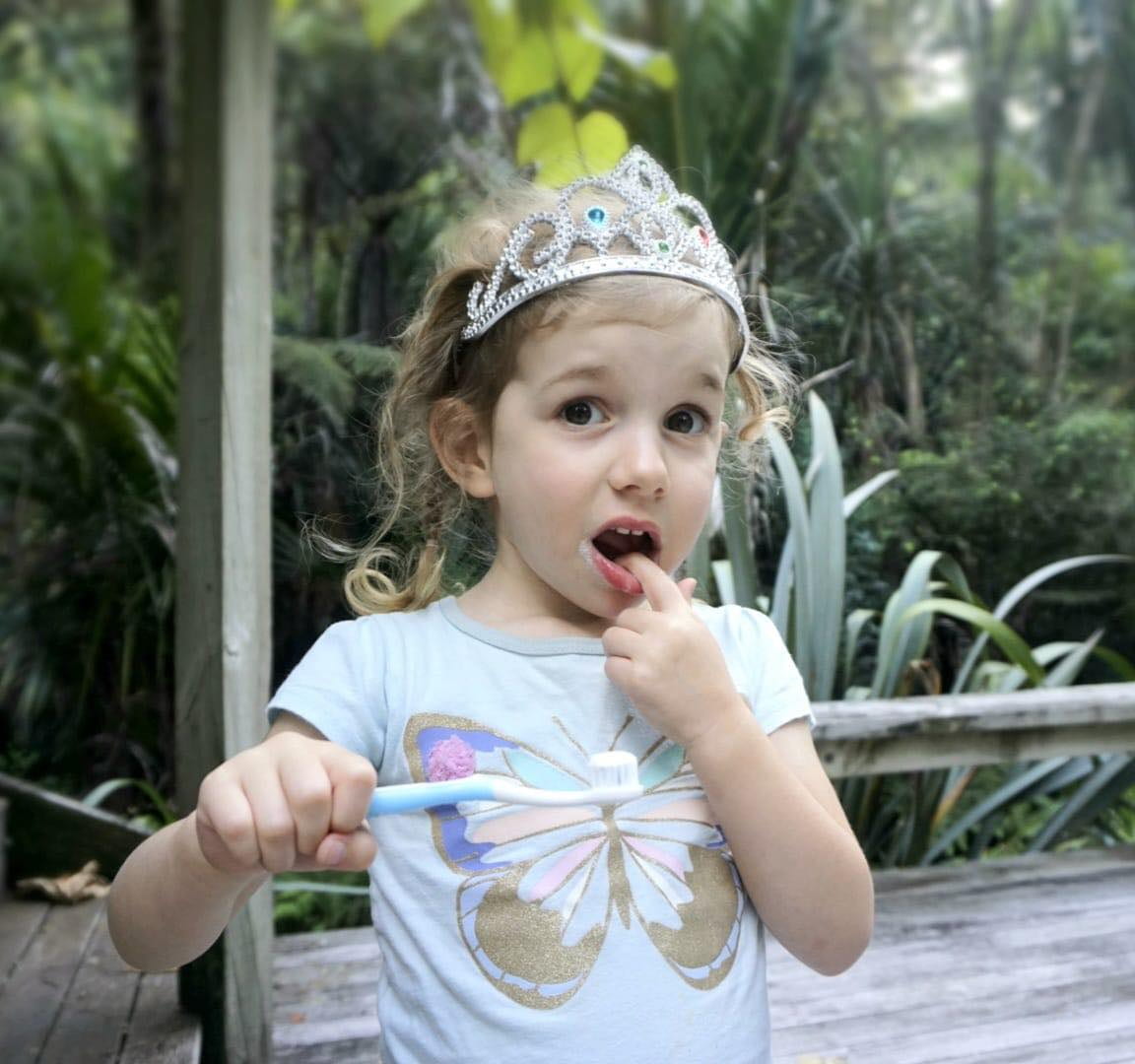 A young girl in a tiara holds a toothbrush with toothpaste. She has her finger in her mouth and a quizzical look on her face