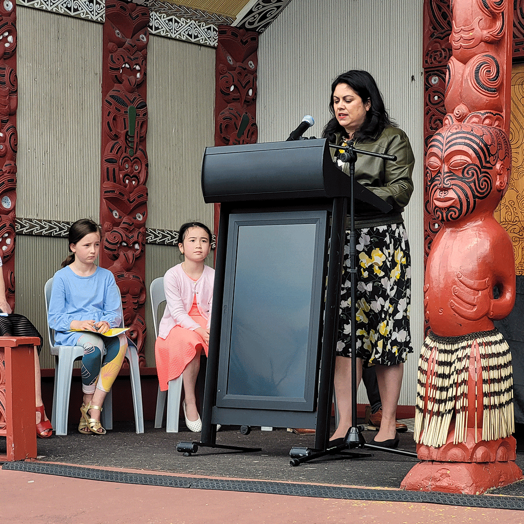 Ayesha Verrall stands at a lectern in front of a marae with two girls sitting on chairs behind her