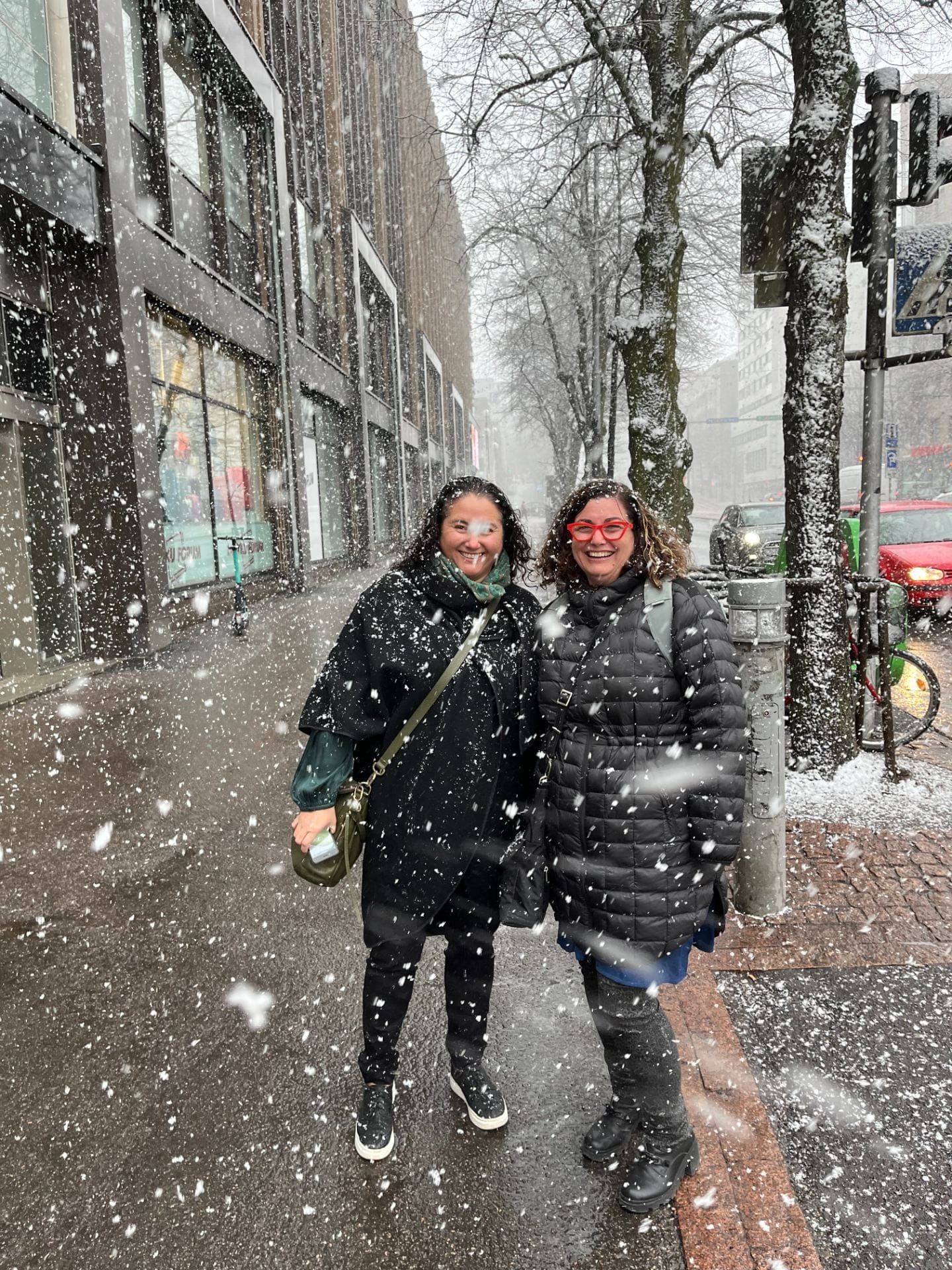 Two women in large black jackets smiling at the camera as snow falls on a sidewalk
